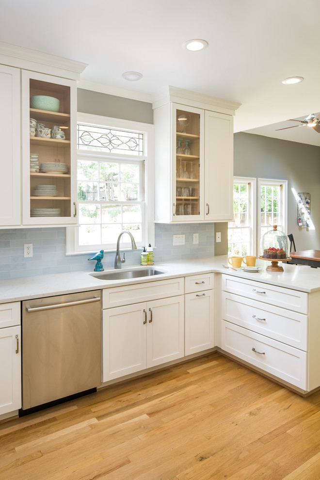 This is an example of a mid-sized transitional l-shaped eat-in kitchen in Atlanta with an undermount sink, recessed-panel cabinets, white cabinets, quartz benchtops, blue splashback, glass tile splashback, stainless steel appliances, light hardwood floors, a peninsula, brown floor and white benchtop.