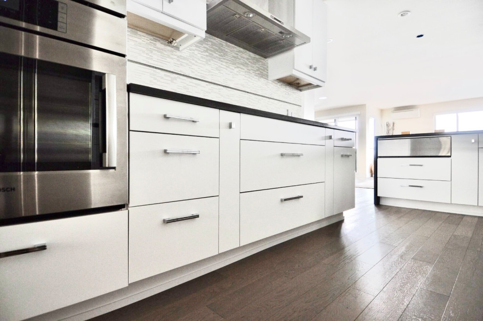 Photo of a large modern l-shaped eat-in kitchen in Seattle with an undermount sink, flat-panel cabinets, white cabinets, quartz benchtops, white splashback, mosaic tile splashback, stainless steel appliances, medium hardwood floors, a peninsula, brown floor and grey benchtop.