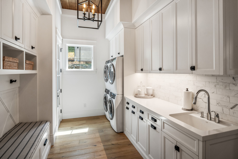 Inspiration for a large transitional galley dedicated laundry room in San Francisco with an undermount sink, shaker cabinets, white cabinets, white splashback, ceramic splashback, white walls, light hardwood floors, a stacked washer and dryer, beige floor, white benchtop and exposed beam.