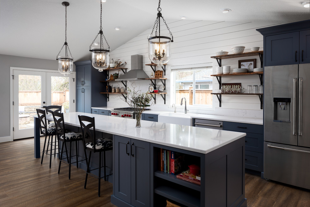 Photo of a large country galley open plan kitchen in Portland with a farmhouse sink, shaker cabinets, blue cabinets, quartz benchtops, white splashback, timber splashback, stainless steel appliances, vinyl floors, with island and brown floor.