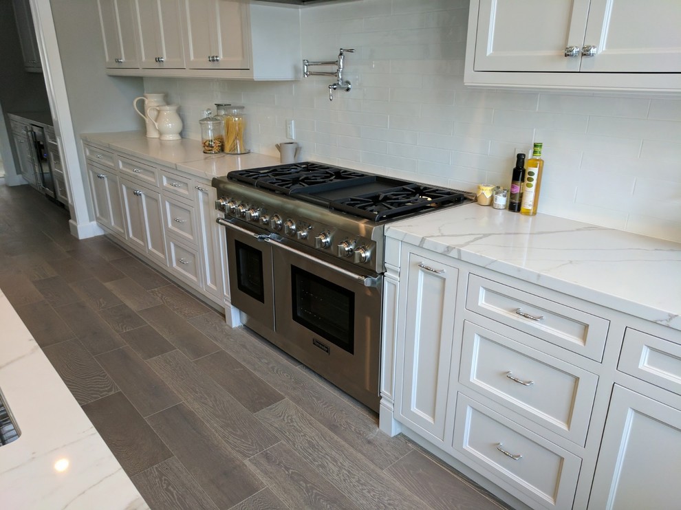 Large beach style u-shaped eat-in kitchen in Orange County with an undermount sink, recessed-panel cabinets, white cabinets, quartz benchtops, white splashback, stone slab splashback, stainless steel appliances, porcelain floors, with island, grey floor, white benchtop and exposed beam.