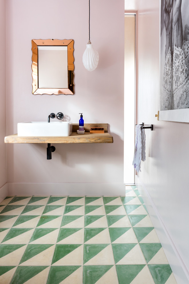 This is an example of a large eclectic powder room in Gloucestershire with pink walls, cement tiles, a wall-mount sink, wood benchtops, green floor and brown benchtops.