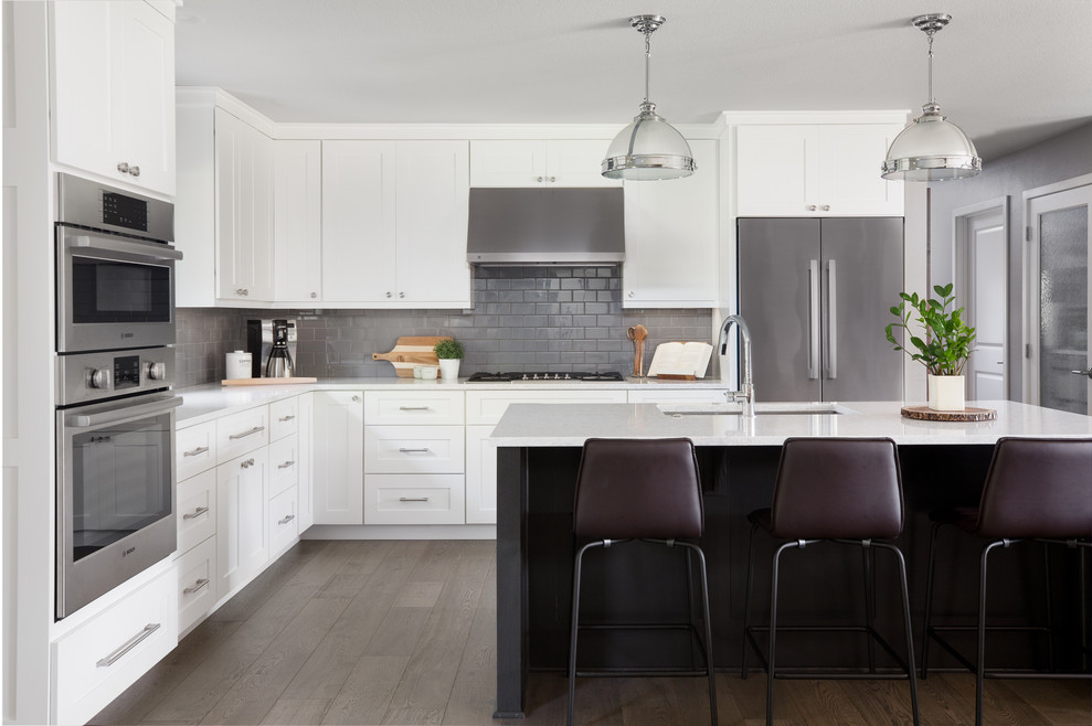 Kitchen - transitional l-shaped medium tone wood floor kitchen idea with an undermount sink, shaker cabinets, white cabinets, gray backsplash, subway tile backsplash, stainless steel appliances, an island and white countertops