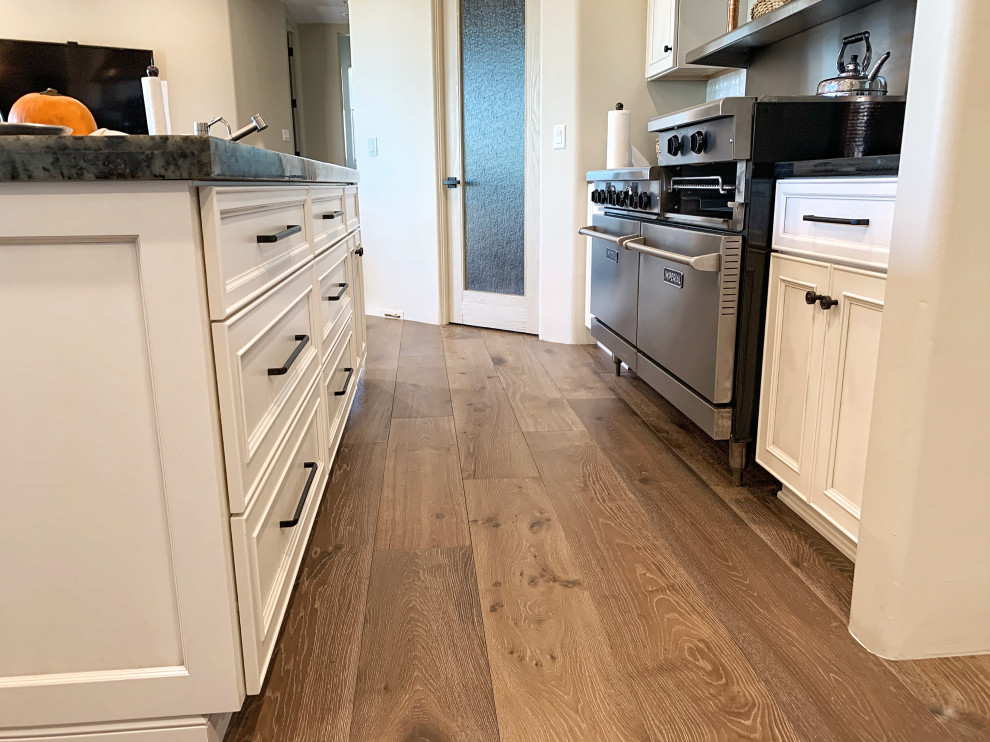 Photo of an expansive transitional u-shaped eat-in kitchen in Phoenix with a farmhouse sink, flat-panel cabinets, white cabinets, quartz benchtops, white splashback, glass tile splashback, stainless steel appliances, light hardwood floors, with island, beige floor and black benchtop.