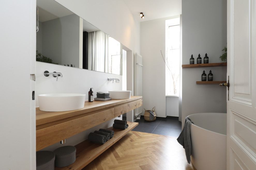 Design ideas for a contemporary bathroom in Frankfurt with flat-panel cabinets, light wood cabinets, a freestanding tub, a curbless shower, a wall-mount toilet, gray tile, light hardwood floors, a vessel sink, solid surface benchtops, grey floor, an open shower, beige benchtops, an enclosed toilet, a double vanity and a floating vanity.