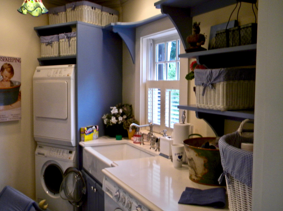 This is an example of a mid-sized traditional galley utility room in Boston with a farmhouse sink, shaker cabinets, blue cabinets, quartz benchtops, white walls, slate floors, a stacked washer and dryer, green floor and white benchtop.