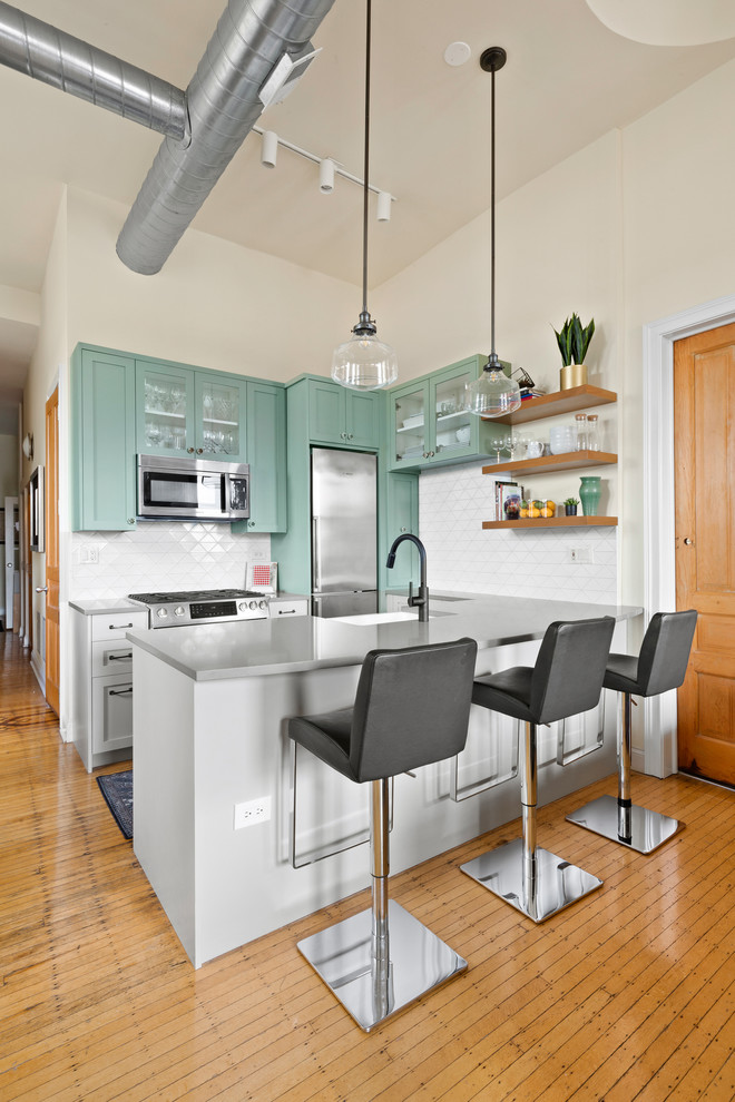 Contemporary u-shaped kitchen in Chicago with a farmhouse sink, shaker cabinets, green cabinets, white splashback, stainless steel appliances, medium hardwood floors, a peninsula, brown floor and grey benchtop.