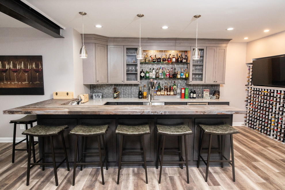 This is an example of a small industrial u-shaped seated home bar in DC Metro with an undermount sink, shaker cabinets, medium wood cabinets, quartz benchtops, grey splashback, metal splashback, vinyl floors, brown floor and grey benchtop.