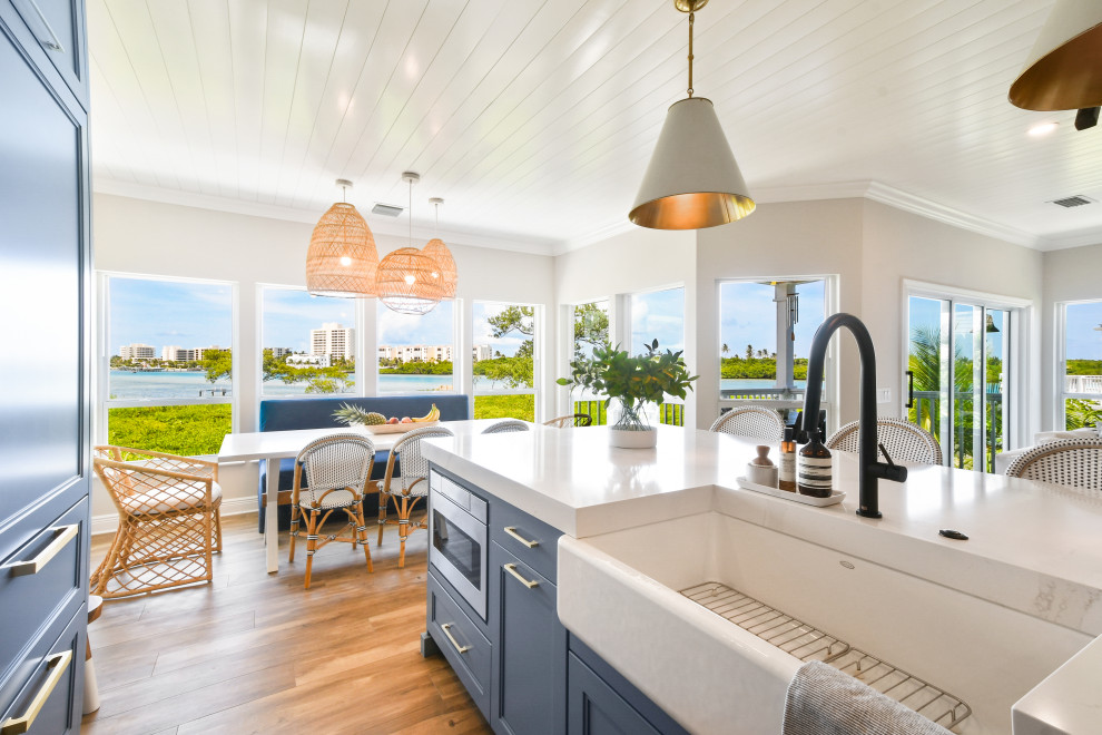Inspiration for a mid-sized beach style l-shaped open plan kitchen in Other with a farmhouse sink, shaker cabinets, blue cabinets, quartz benchtops, white splashback, porcelain splashback, black appliances, porcelain floors, with island, brown floor, white benchtop and timber.