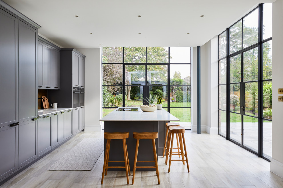This is an example of a traditional kitchen/diner in Surrey with an integrated sink, shaker cabinets, grey cabinets, marble worktops, metallic splashback, black appliances, light hardwood flooring, an island, grey floors and white worktops.