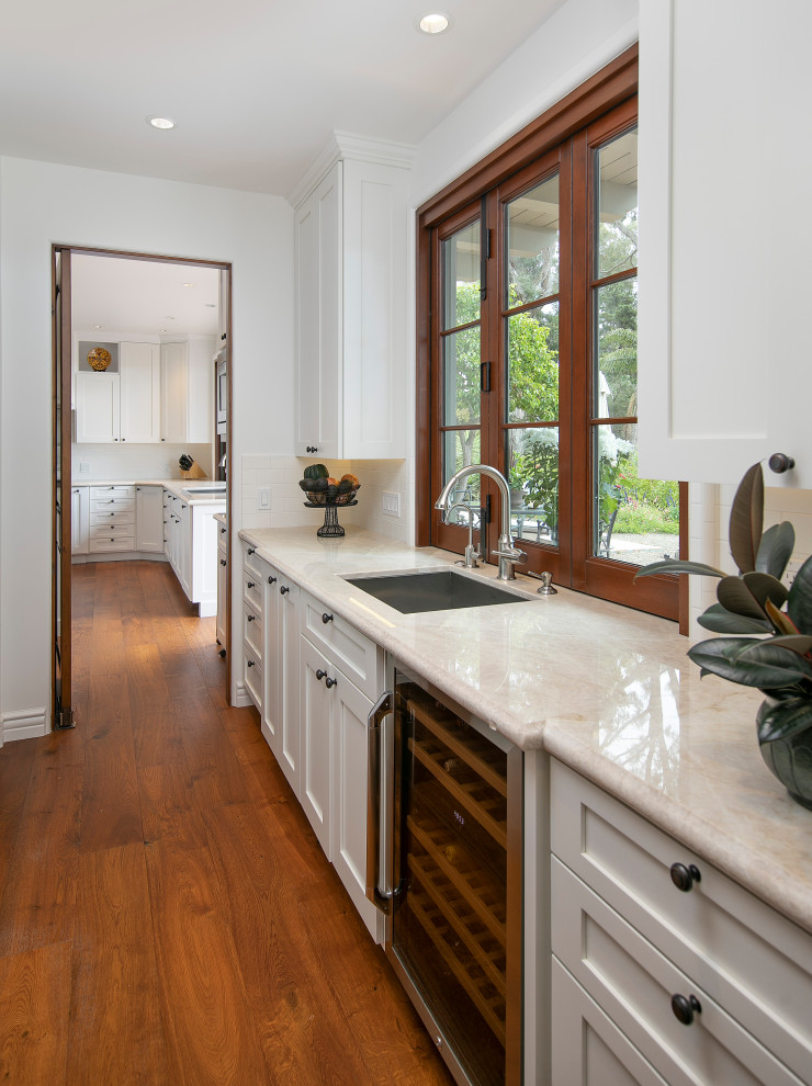 Inspiration for a mid-sized mediterranean u-shaped medium tone wood floor and brown floor home bar remodel in Santa Barbara with recessed-panel cabinets, white cabinets, marble countertops, white backsplash, ceramic backsplash and white countertops