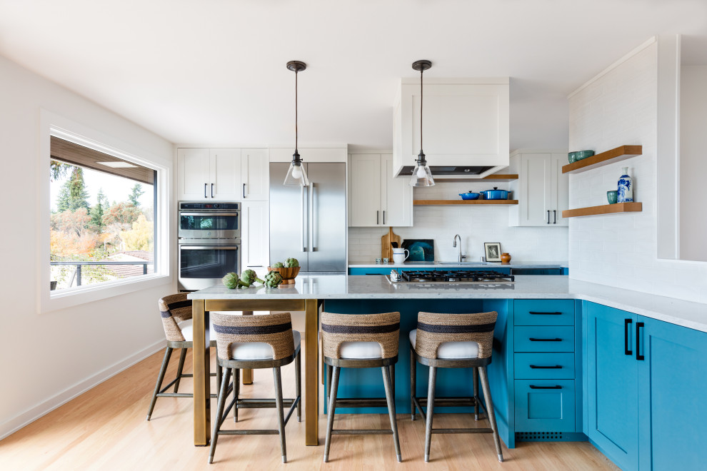 This is an example of a transitional kitchen in Seattle with an undermount sink, shaker cabinets, blue cabinets, quartz benchtops, white splashback, ceramic splashback, stainless steel appliances, light hardwood floors, a peninsula and white benchtop.