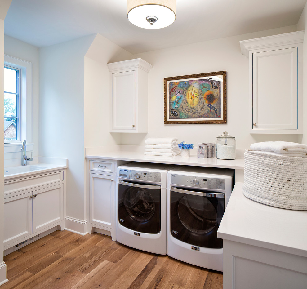 Design ideas for a mid-sized traditional u-shaped dedicated laundry room in Minneapolis with white cabinets, quartz benchtops, white walls, light hardwood floors, a side-by-side washer and dryer, an undermount sink, recessed-panel cabinets and white benchtop.