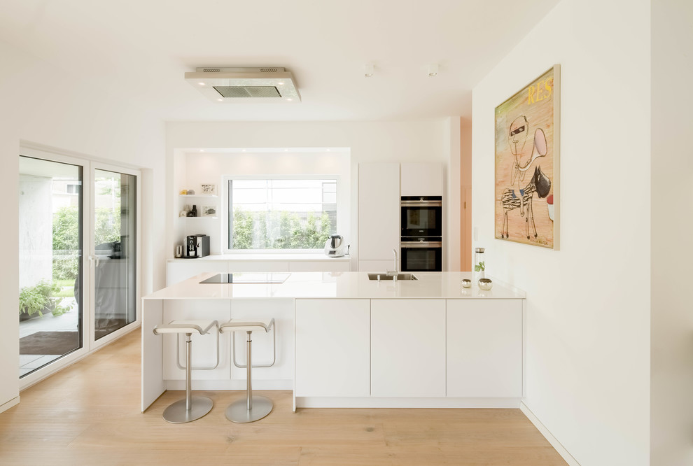 Inspiration for a mid-sized contemporary galley open plan kitchen in Dusseldorf with a drop-in sink, flat-panel cabinets, white cabinets, white splashback, stainless steel appliances, light hardwood floors and with island.