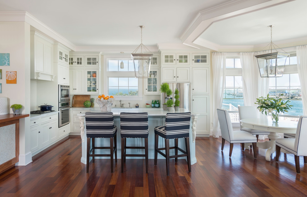 Photo of a mid-sized beach style l-shaped eat-in kitchen in Providence with a farmhouse sink, recessed-panel cabinets, white cabinets, marble benchtops, white splashback, ceramic splashback, stainless steel appliances, medium hardwood floors and with island.