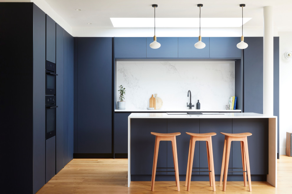 Inspiration for a large contemporary l-shaped open plan kitchen in London with an undermount sink, flat-panel cabinets, blue cabinets, quartzite benchtops, white splashback, engineered quartz splashback, black appliances, medium hardwood floors, with island and white benchtop.