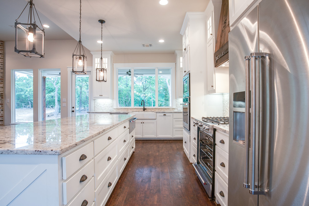 Design ideas for a large country l-shaped open plan kitchen in Dallas with a farmhouse sink, shaker cabinets, white cabinets, granite benchtops, white splashback, matchstick tile splashback, stainless steel appliances, medium hardwood floors, with island and brown floor.