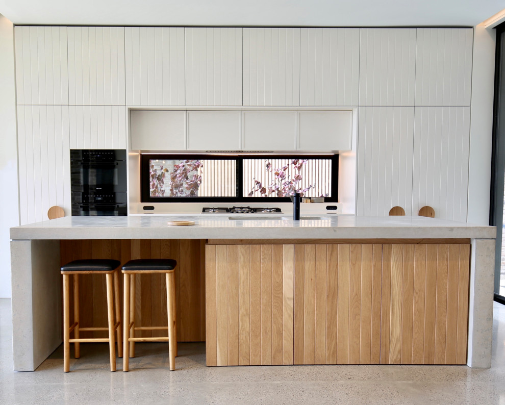 Design ideas for a beach style galley open plan kitchen in Sydney with an undermount sink, shaker cabinets, light wood cabinets, concrete benchtops, window splashback, black appliances, concrete floors, with island, grey floor and white benchtop.