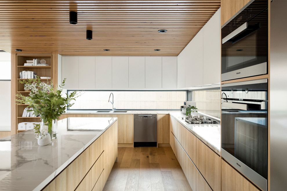 This is an example of a large contemporary l-shaped open plan kitchen in Melbourne with an undermount sink, flat-panel cabinets, light wood cabinets, marble benchtops, window splashback, black appliances, light hardwood floors, with island, brown floor, white benchtop and timber.