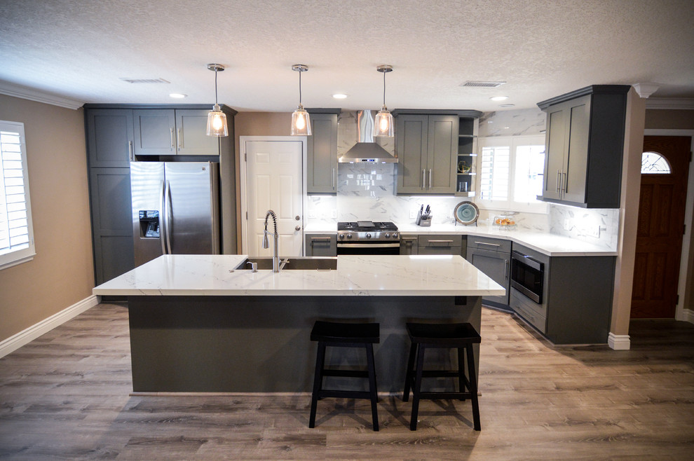 This is an example of an expansive contemporary l-shaped eat-in kitchen in Orange County with a double-bowl sink, recessed-panel cabinets, grey cabinets, quartz benchtops, grey splashback, porcelain splashback, stainless steel appliances, laminate floors, with island, beige floor and grey benchtop.