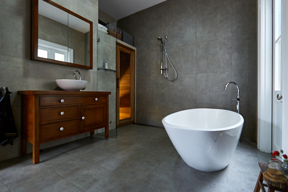 This is an example of a mid-sized traditional 3/4 bathroom in Sydney with furniture-like cabinets, brown cabinets, a freestanding tub, an open shower, a wall-mount toilet, gray tile, ceramic tile, grey walls, ceramic floors, a pedestal sink, wood benchtops, grey floor, an open shower, brown benchtops, an enclosed toilet, a single vanity and a freestanding vanity.