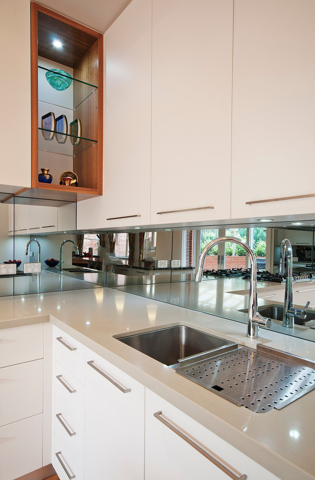 This is an example of a mid-sized modern l-shaped open plan kitchen in Melbourne with an undermount sink, flat-panel cabinets, white cabinets, metallic splashback, glass sheet splashback, stainless steel appliances, medium hardwood floors, with island and quartz benchtops.