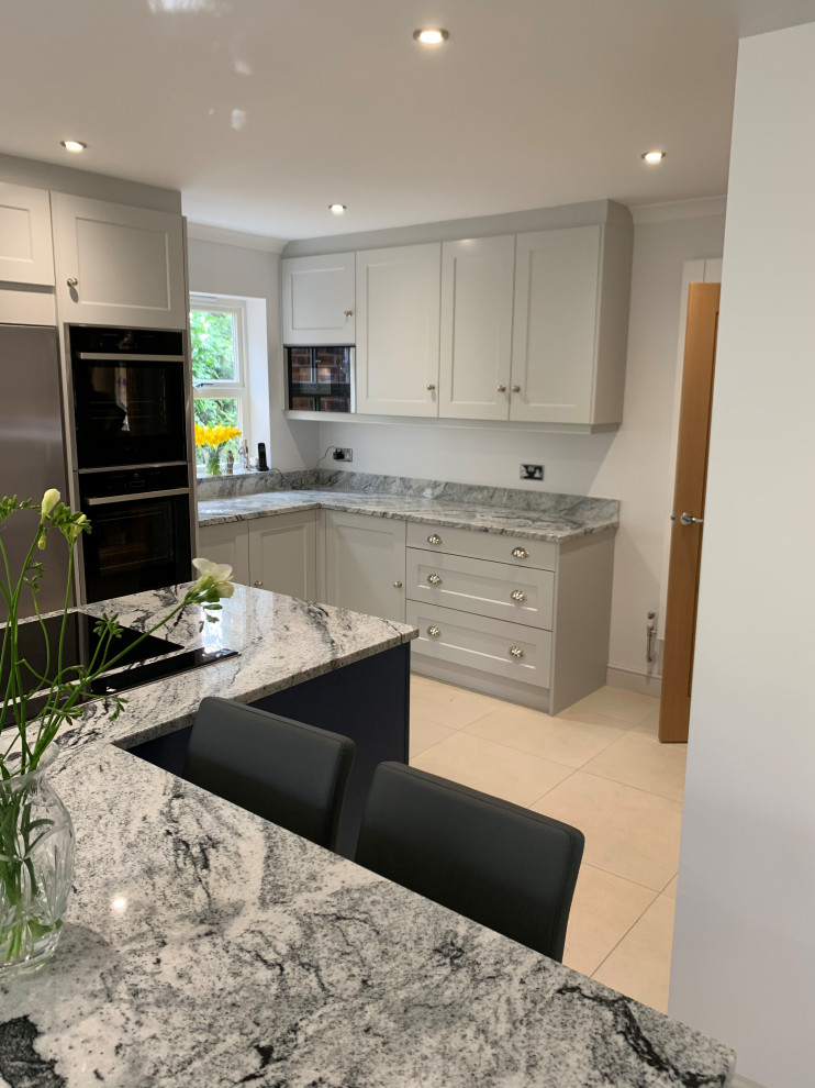 Photo of a large l-shaped open plan kitchen in Sussex with a farmhouse sink, shaker cabinets, grey cabinets, granite benchtops, grey splashback, granite splashback, black appliances, porcelain floors, with island, beige floor and grey benchtop.