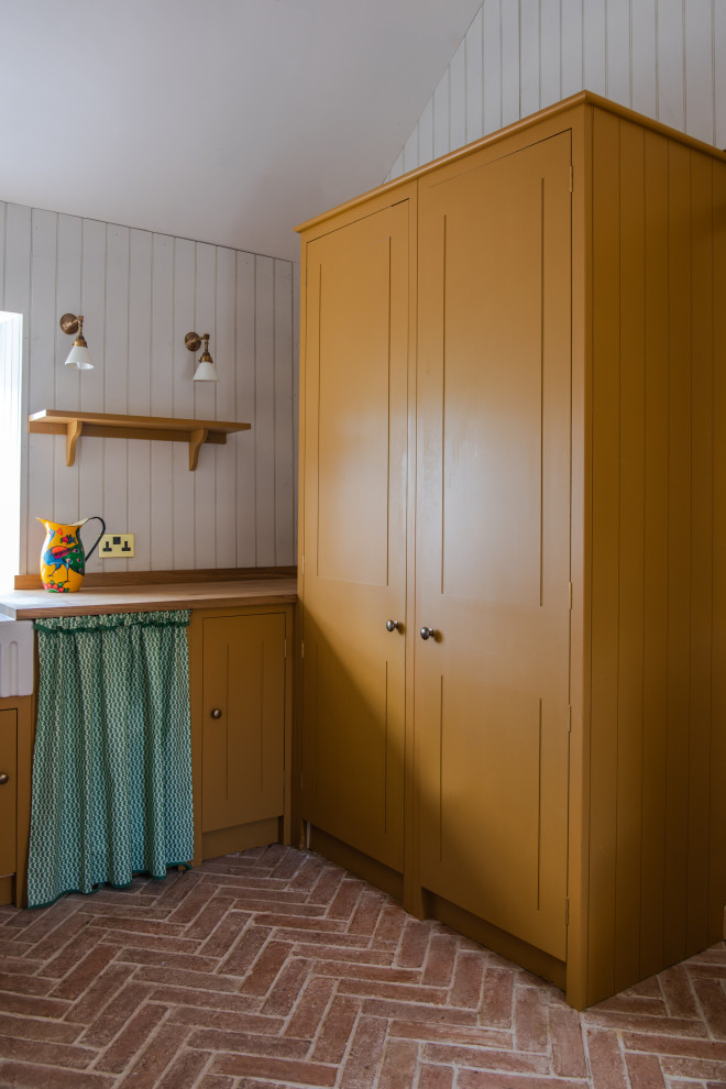 This is an example of a small modern l-shaped utility room in London with a farmhouse sink, beaded inset cabinets, yellow cabinets, wood benchtops, white walls, brick floors, brown floor and brown benchtop.