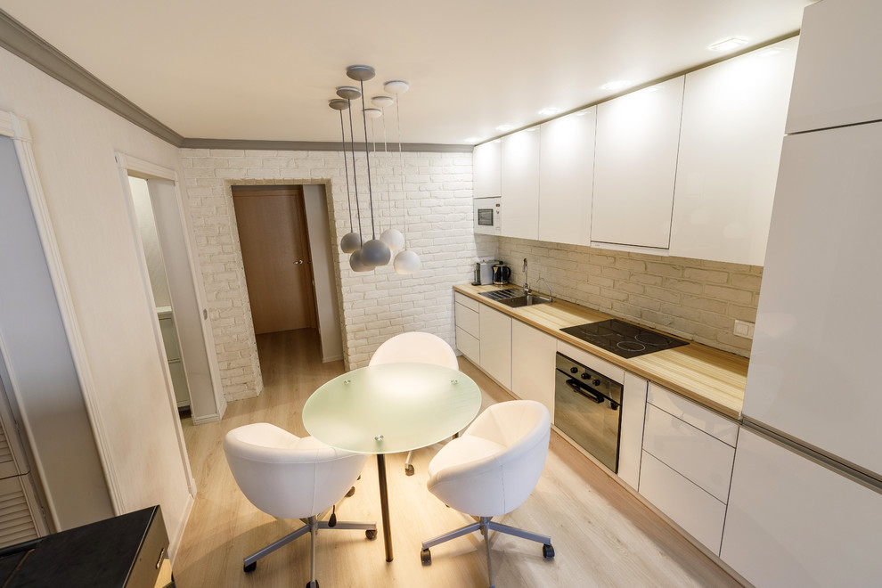 Photo of a mid-sized contemporary single-wall eat-in kitchen in Saint Petersburg with an undermount sink, flat-panel cabinets, white cabinets, wood benchtops, white splashback, brick splashback, stainless steel appliances, light hardwood floors and grey floor.