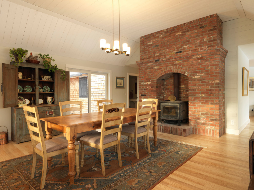 Inspiration for a mid-sized country dining room in Burlington with grey walls, light hardwood floors, a wood stove, a brick fireplace surround, brown floor and timber.