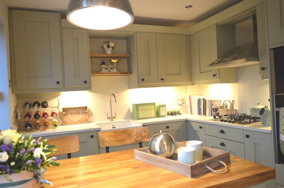 Photo of a small traditional u-shaped separate kitchen in Other with a farmhouse sink, shaker cabinets, green cabinets, quartzite benchtops, white splashback, stone slab splashback, panelled appliances, laminate floors, a peninsula, brown floor and white benchtop.