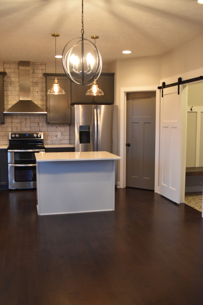 Example of a mid-sized eclectic l-shaped medium tone wood floor and brown floor open concept kitchen design with a farmhouse sink, raised-panel cabinets, gray cabinets, brick backsplash, stainless steel appliances and an island