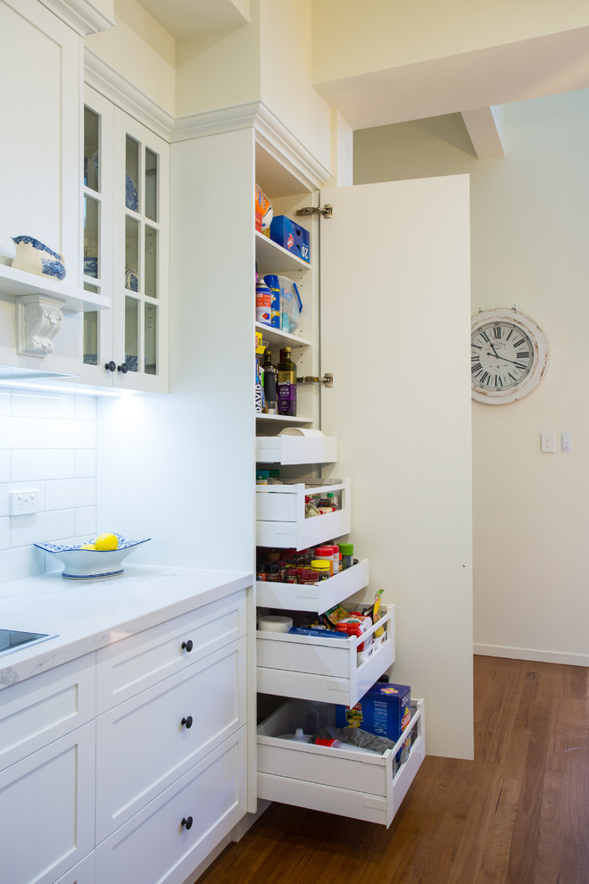 This is an example of a large traditional galley kitchen pantry in Brisbane with a farmhouse sink, shaker cabinets, white cabinets, quartz benchtops, white splashback, subway tile splashback, white appliances, medium hardwood floors, with island, brown floor and white benchtop.