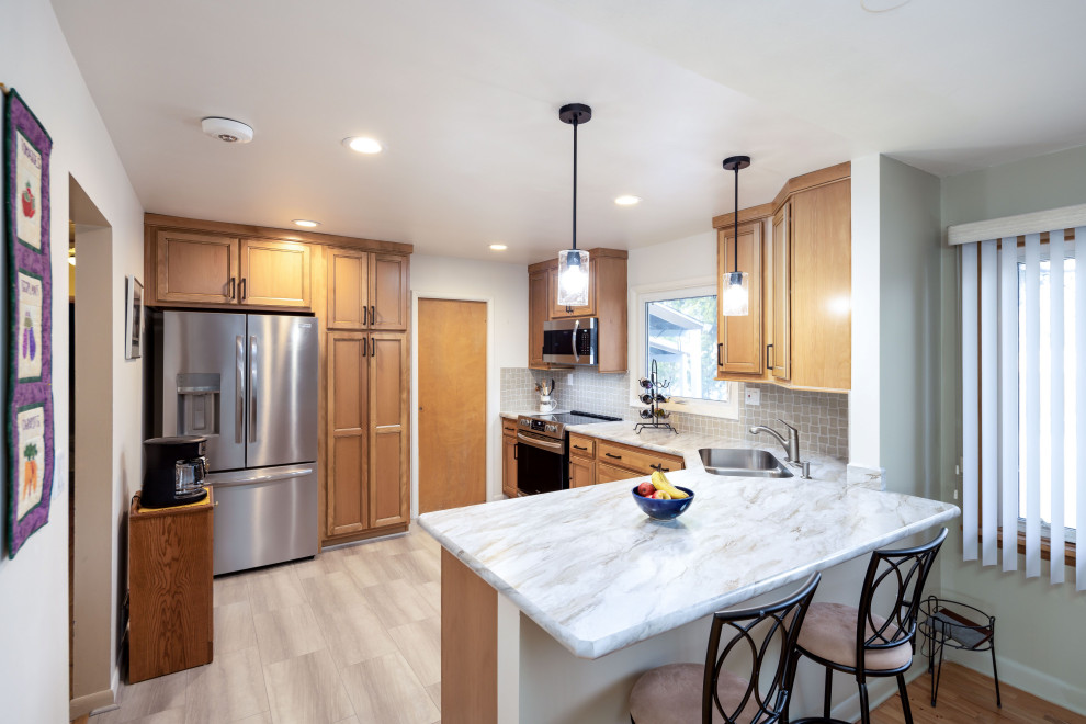 Mid-sized transitional l-shaped light wood floor and gray floor eat-in kitchen photo with an integrated sink, recessed-panel cabinets, light wood cabinets, laminate countertops, gray backsplash, porcelain backsplash, stainless steel appliances, a peninsula and white countertops