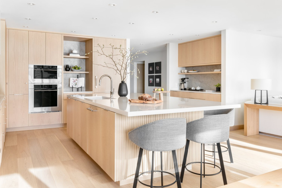 This is an example of a mid-sized modern l-shaped eat-in kitchen in Vancouver with a double-bowl sink, flat-panel cabinets, light wood cabinets, solid surface benchtops, grey splashback, limestone splashback, panelled appliances, light hardwood floors, with island, beige floor and white benchtop.