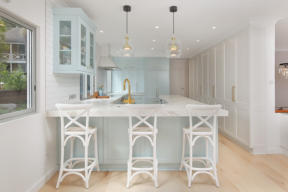 Photo of a large transitional u-shaped eat-in kitchen in Sydney with a farmhouse sink, shaker cabinets, blue cabinets, quartz benchtops, white splashback, subway tile splashback, panelled appliances, light hardwood floors, beige floor and grey benchtop.