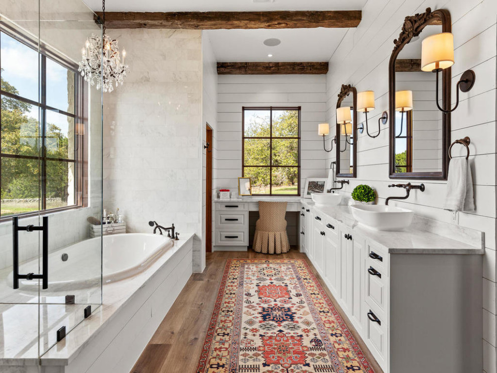 Country bathroom in Austin with raised-panel cabinets, white cabinets, a drop-in tub, gray tile, marble, white walls, medium hardwood floors, a vessel sink, marble benchtops, beige floor, a hinged shower door, grey benchtops, a double vanity, a built-in vanity, exposed beam and planked wall panelling.