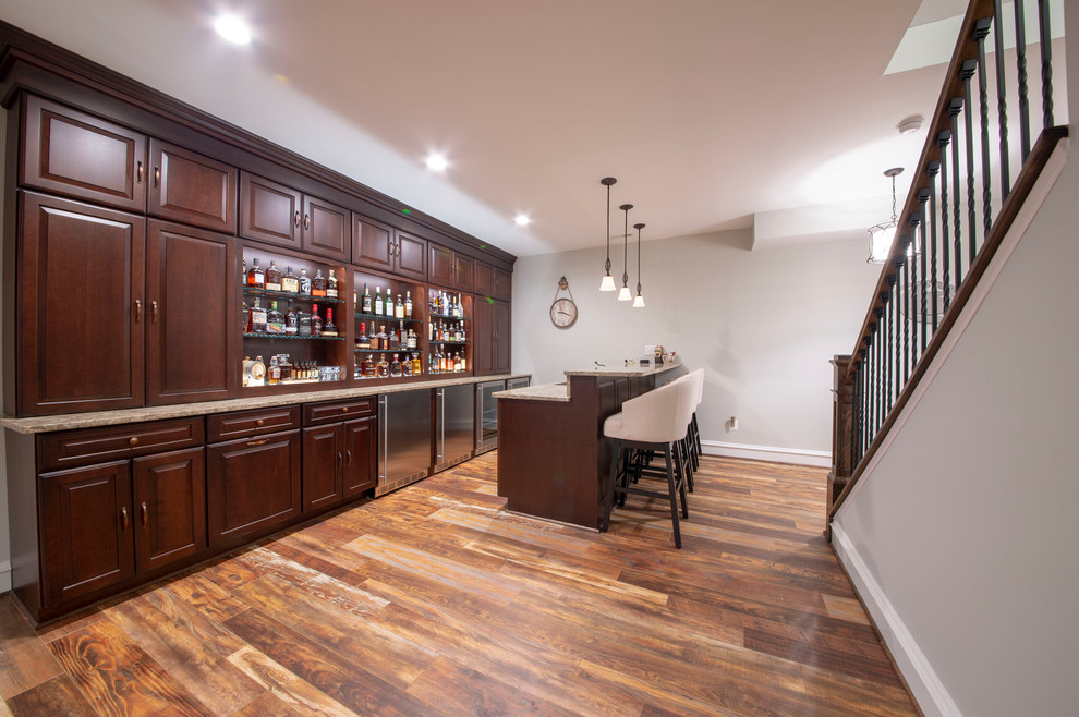 Photo of an expansive arts and crafts single-wall seated home bar in DC Metro with an undermount sink, raised-panel cabinets, dark wood cabinets, granite benchtops, medium hardwood floors, brown floor and multi-coloured benchtop.
