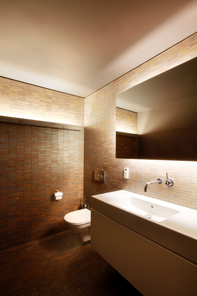 This is an example of a mid-sized modern powder room in Cologne with flat-panel cabinets, white cabinets, a wall-mount toilet, brown tile, matchstick tile, brown walls and an integrated sink.