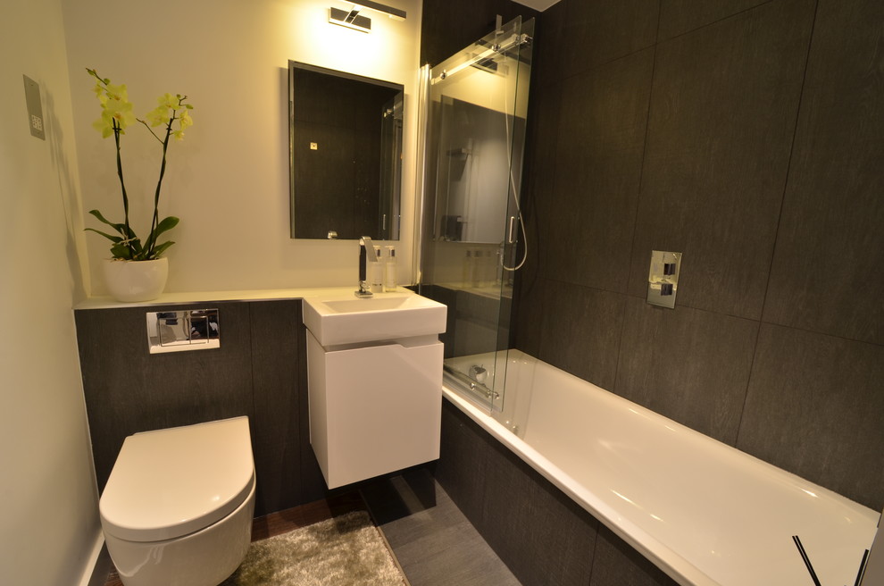 This is an example of a mid-sized contemporary kids bathroom in London with flat-panel cabinets, white cabinets, a drop-in tub, a shower/bathtub combo, a wall-mount toilet, brown tile, ceramic tile, beige walls, dark hardwood floors, a wall-mount sink and glass benchtops.