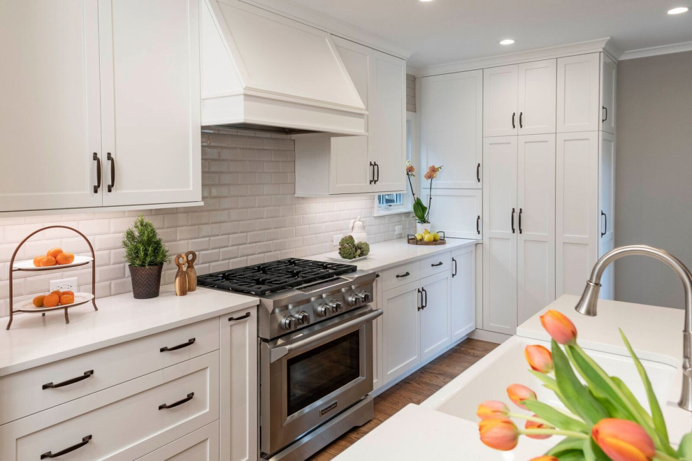 Mid-sized transitional u-shaped eat-in kitchen in Indianapolis with a farmhouse sink, shaker cabinets, white cabinets, quartz benchtops, white splashback, ceramic splashback, stainless steel appliances, medium hardwood floors, with island, brown floor and white benchtop.