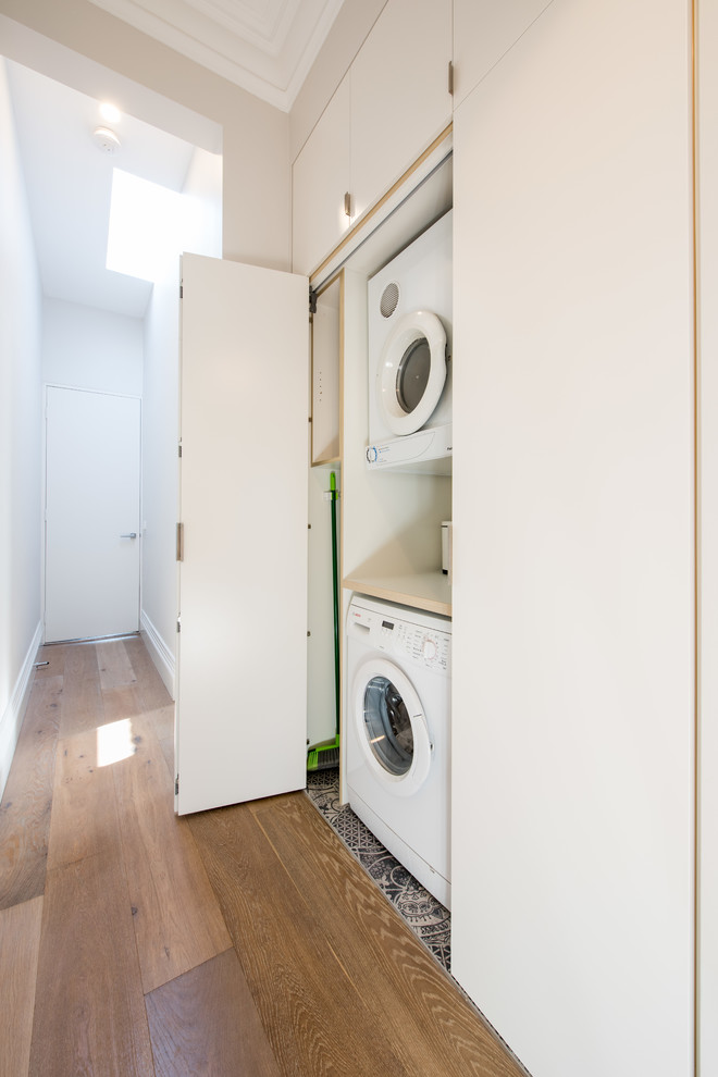 This is an example of a transitional single-wall laundry cupboard in Melbourne with flat-panel cabinets, white cabinets, laminate benchtops, a drop-in sink, porcelain floors and a stacked washer and dryer.