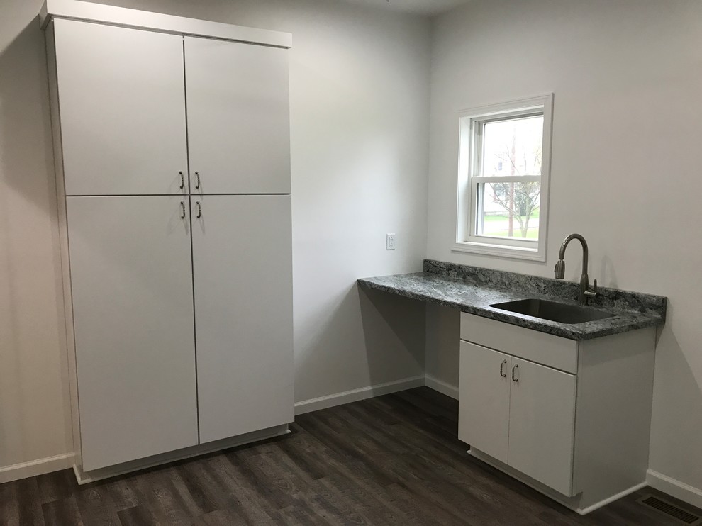 This is an example of a mid-sized country l-shaped laundry room in Other with a farmhouse sink, flat-panel cabinets, white cabinets, quartz benchtops, grey splashback, cement tile splashback and brown floor.