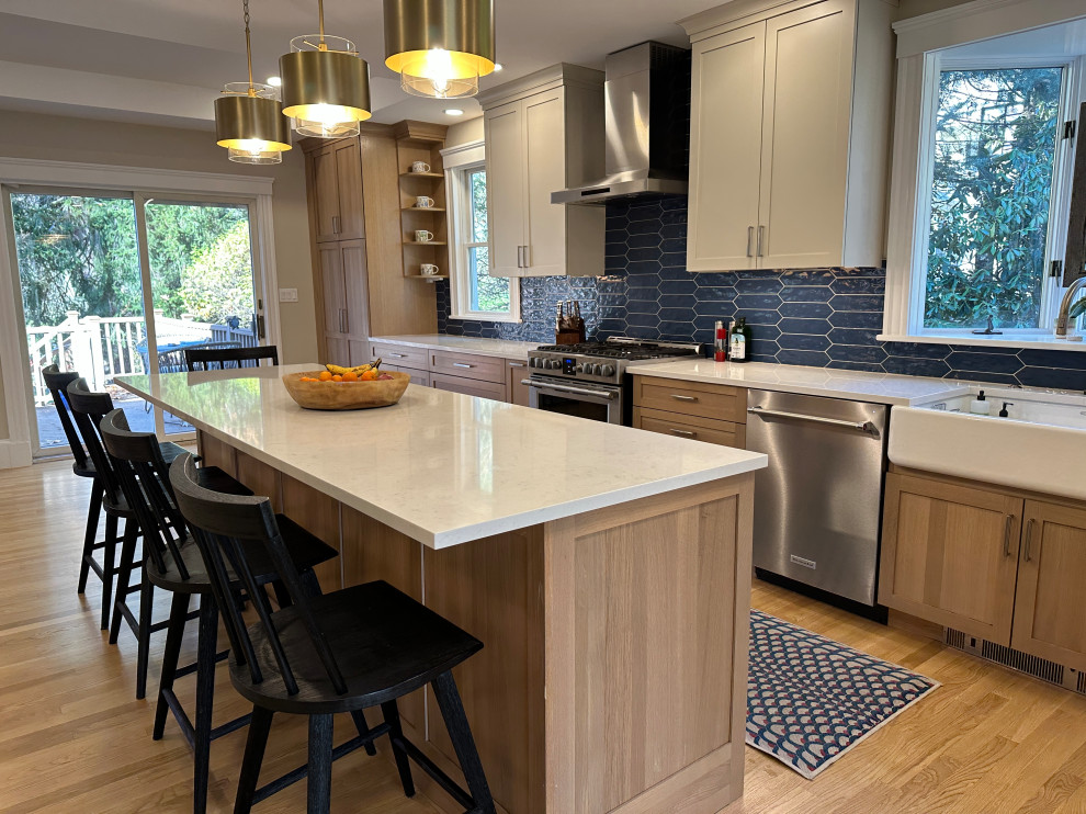 Design ideas for a large country l-shaped eat-in kitchen in Boston with a farmhouse sink, shaker cabinets, light wood cabinets, quartz benchtops, blue splashback, cement tile splashback, stainless steel appliances, light hardwood floors, with island and white benchtop.
