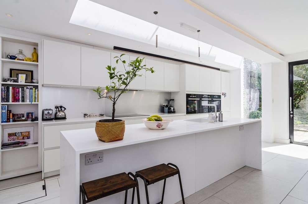 This is an example of a large single-wall eat-in kitchen in London with an integrated sink, flat-panel cabinets, white cabinets, solid surface benchtops, white splashback, stainless steel appliances, porcelain floors and a peninsula.
