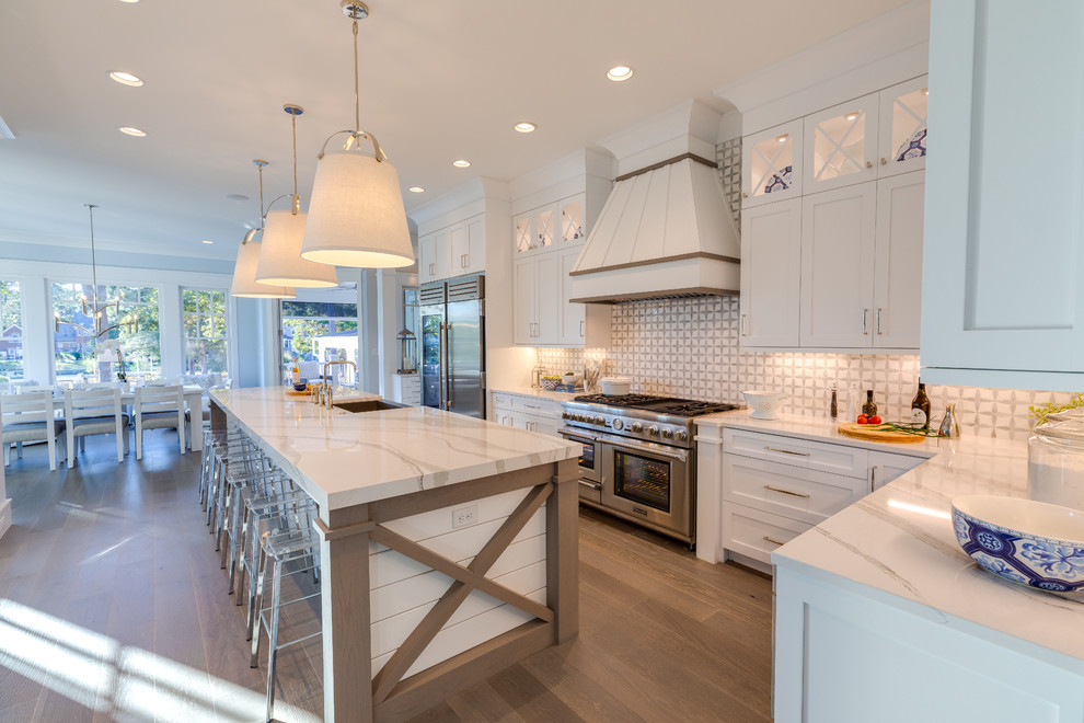 Photo of an expansive beach style l-shaped eat-in kitchen in Other with a farmhouse sink, recessed-panel cabinets, white cabinets, quartz benchtops, white splashback, marble splashback, stainless steel appliances, medium hardwood floors, with island, grey floor and white benchtop.