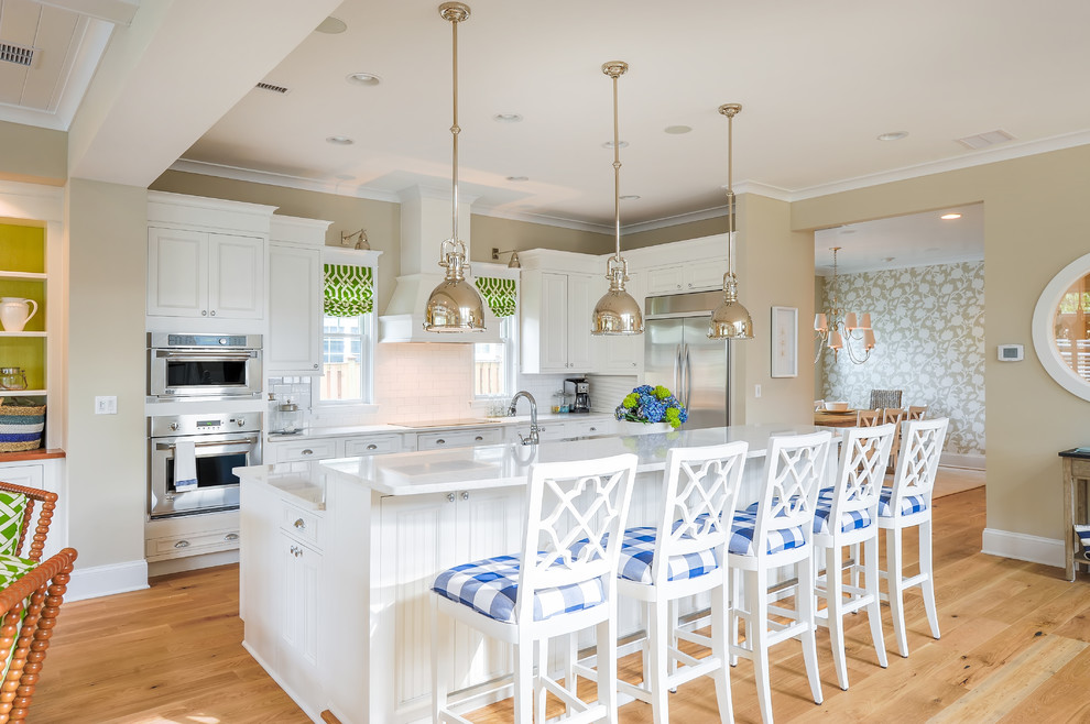 This is an example of a mid-sized beach style open plan kitchen in Philadelphia with recessed-panel cabinets, white cabinets, quartz benchtops, white splashback, ceramic splashback, stainless steel appliances, light hardwood floors and with island.