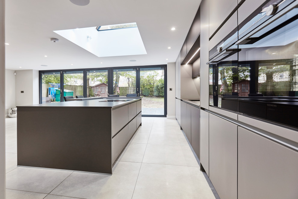 This is an example of a mid-sized contemporary single-wall open plan kitchen in Other with an integrated sink, flat-panel cabinets, grey cabinets, quartzite benchtops, metallic splashback, mirror splashback, black appliances, porcelain floors, with island, grey floor, grey benchtop and vaulted.
