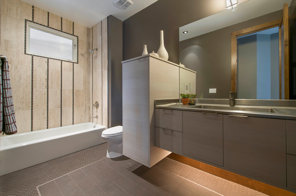 This is an example of a small contemporary bathroom in Denver with an integrated sink, flat-panel cabinets, light wood cabinets, stainless steel benchtops, an alcove tub, a shower/bathtub combo, a one-piece toilet, beige tile, stone tile, brown walls and porcelain floors.