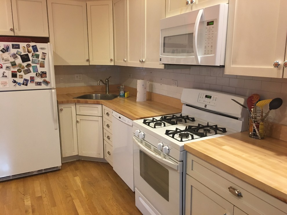 This is an example of a mid-sized country l-shaped eat-in kitchen in Chicago with an undermount sink, shaker cabinets, white cabinets, wood benchtops, white splashback, subway tile splashback, white appliances, light hardwood floors, no island and brown floor.
